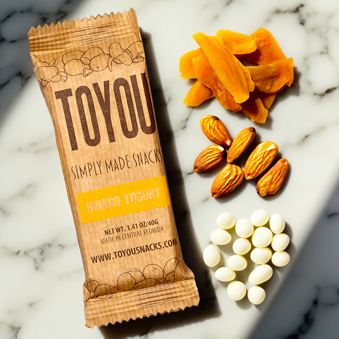 Top view: One Mango Yogurt ToYou snack bar on white marble with piles of dried mango slices, almonds, and yogurt chips next to it. 