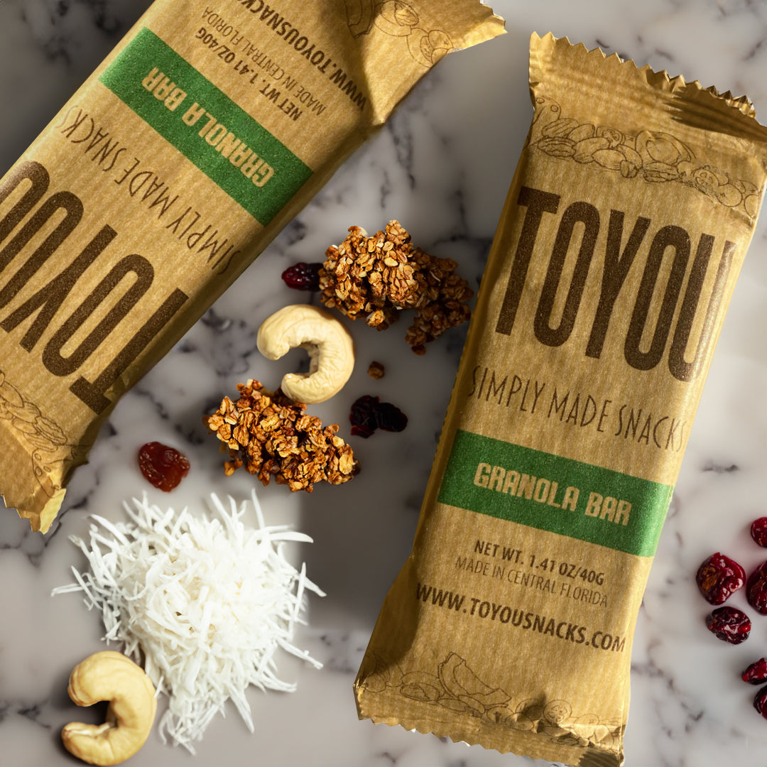Top view: Two Granola Bar ToYou snack bars on white marble with granola chunks, shredded coconut, cashews, and dried cranberries scattered around. 