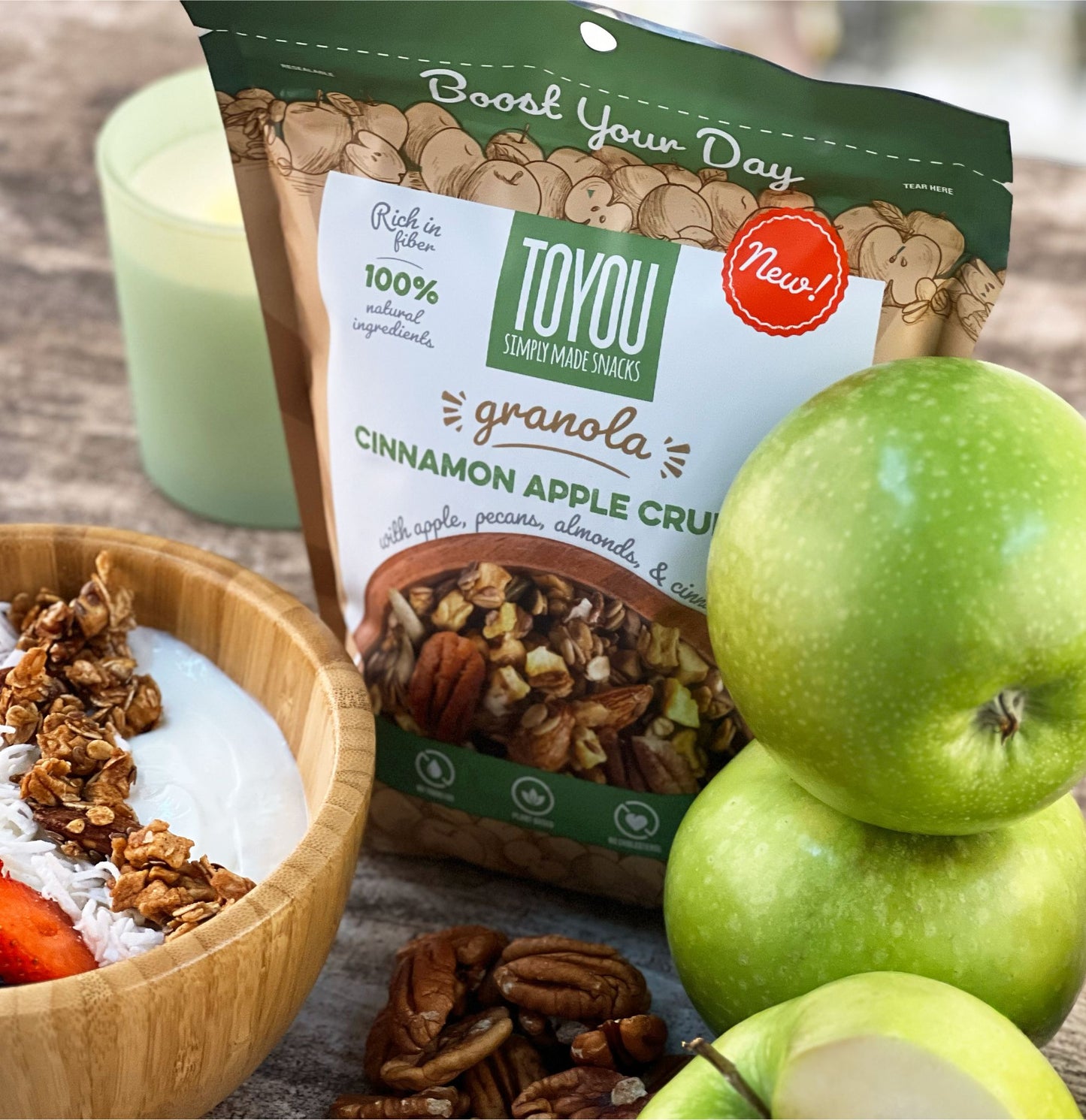 Isometric view: Green granola pouch on wood surface (Artwork: top view photo, wooden bowl with yogurt, strawberries, shredded coconut, Cinnamon Apple Crunch ToYou Granola). Behind pouch: Green candle. Front right: Two green apples, sliced green apple half