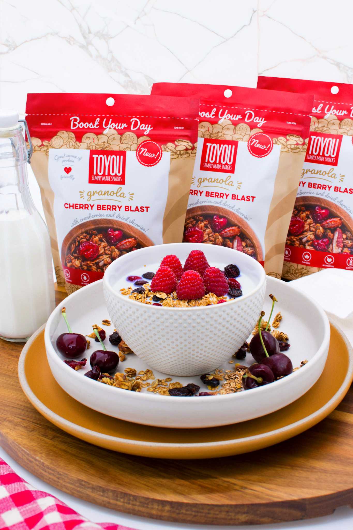 Front view: 3 red Cherry Berry Blast ToYou Granola pouches next milk glass bottle, behind white bowl with yogurt, granola and cherries. 