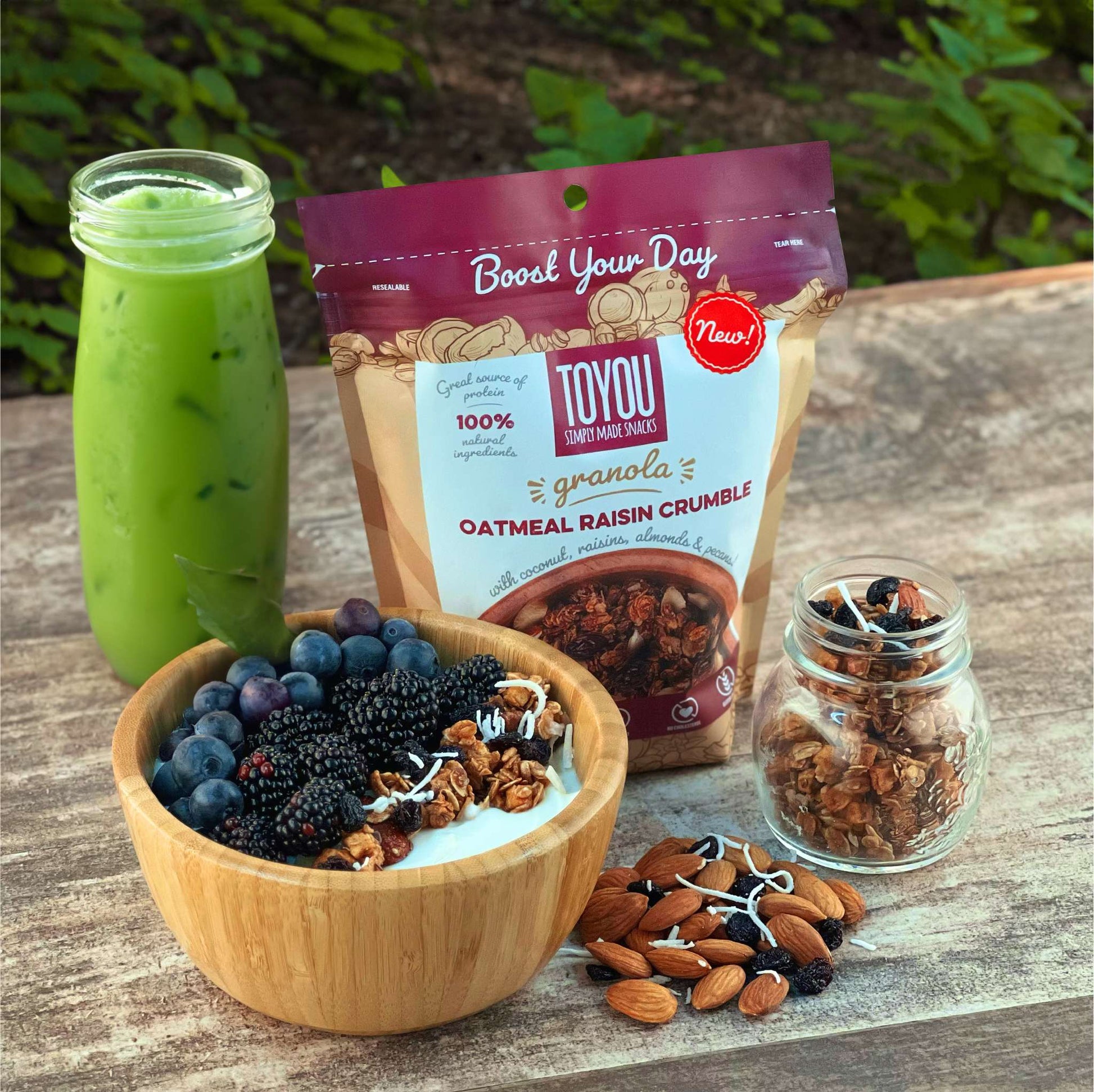 Isometric view: Purple granola pouch on wood surface with forest like background (Artwork: Wooden bowl with yogurt, blueberries, blackberries, shredded coconut, Oatmeal Raisin Crumble ToYou Granola) Behind: Tall clear glass with green juice. Front right: 